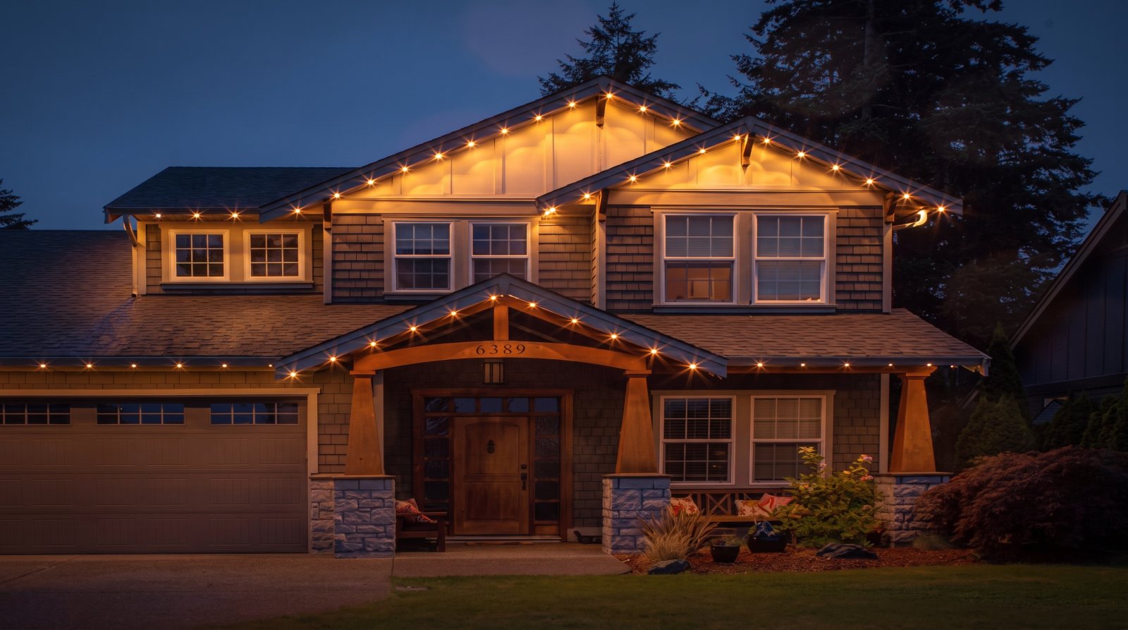 Two-storey craftsman home at dusk with warm white permanent lighting tracing every roofline, gable and the porch