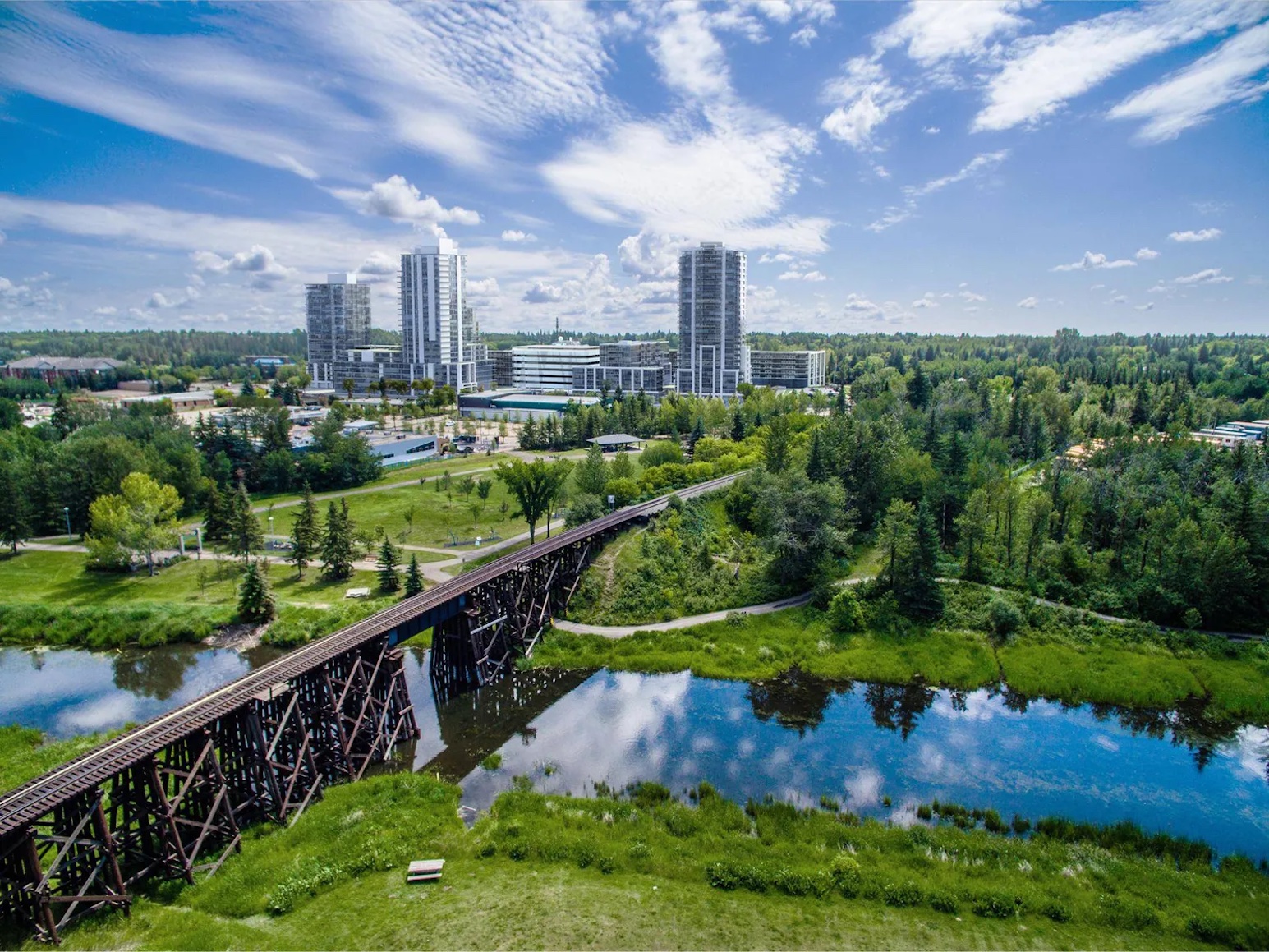 Aerial view of St. Albert, Alberta in summer — the historic railway trestle bridge over the Sturgeon River with the downtown skyline